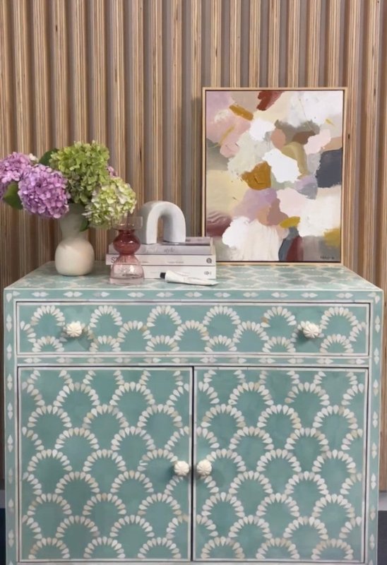 Teal blue bone inlay scalloped cabinet with one drawer and two doors with white ceramic knobs, styled with hydrangea flowers, books, sculptures and abstract canvas art on slatted wood wall