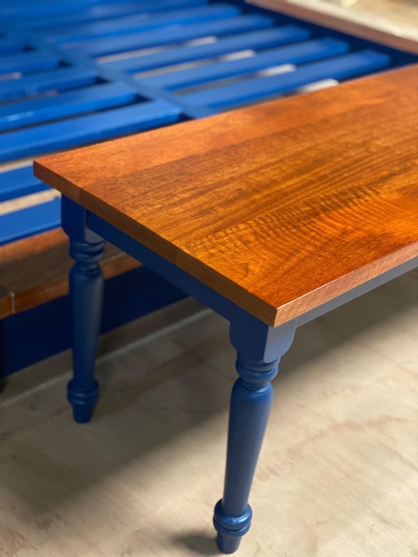Turned leg seat bench with blue wooden supports, corner detail view showing the warm wood grain and seat edge.