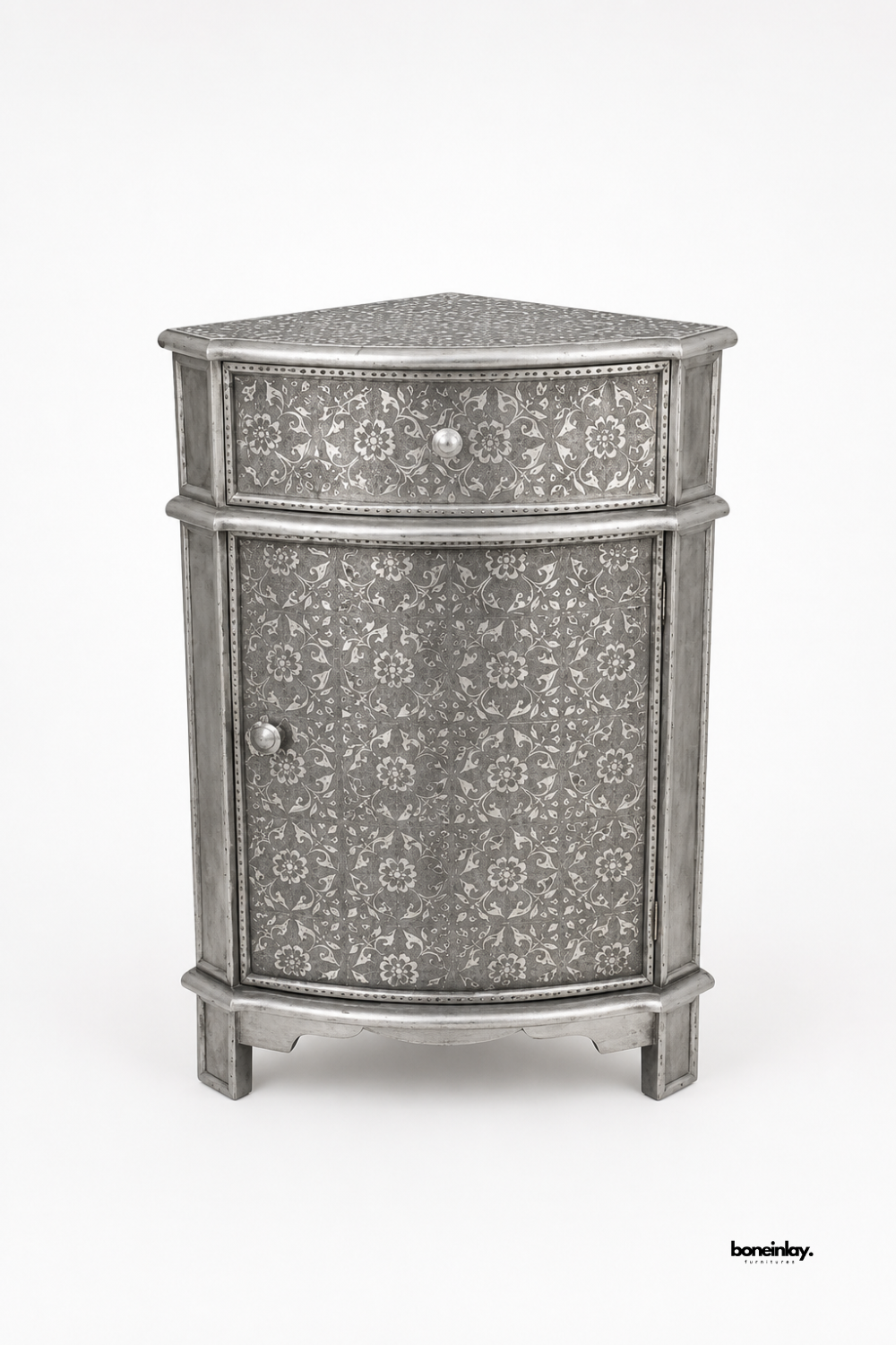 Artisan-crafted corner storage cabinet wrapped in hand-embossed silver sheet metal with traditional botanical motifs and beaded trim detailing. Showcased against a clean studio background, this space-saving triangular accent chest features a top drawer, a lower cabinet with knob hardware, and elegant tapered feet for distinctive home decor.
