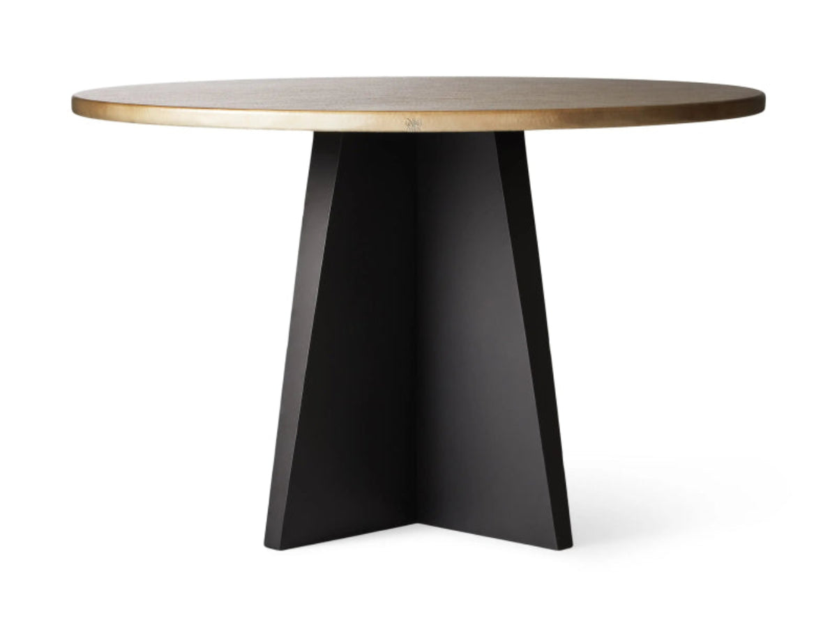 Round brushed brass top dining table with a matte black crossed four-panel pedestal base against a gray studio background.