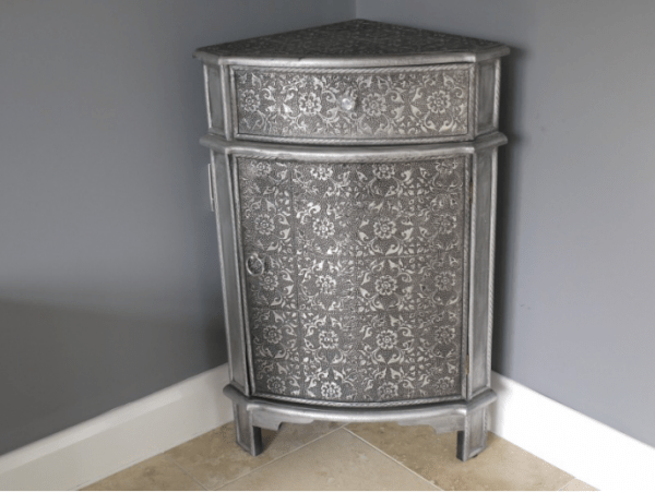 White metal corner cabinet chest featuring hand-embossed silver floral cladding, a crystal drawer knob, and a ring pull on short wooden feet.