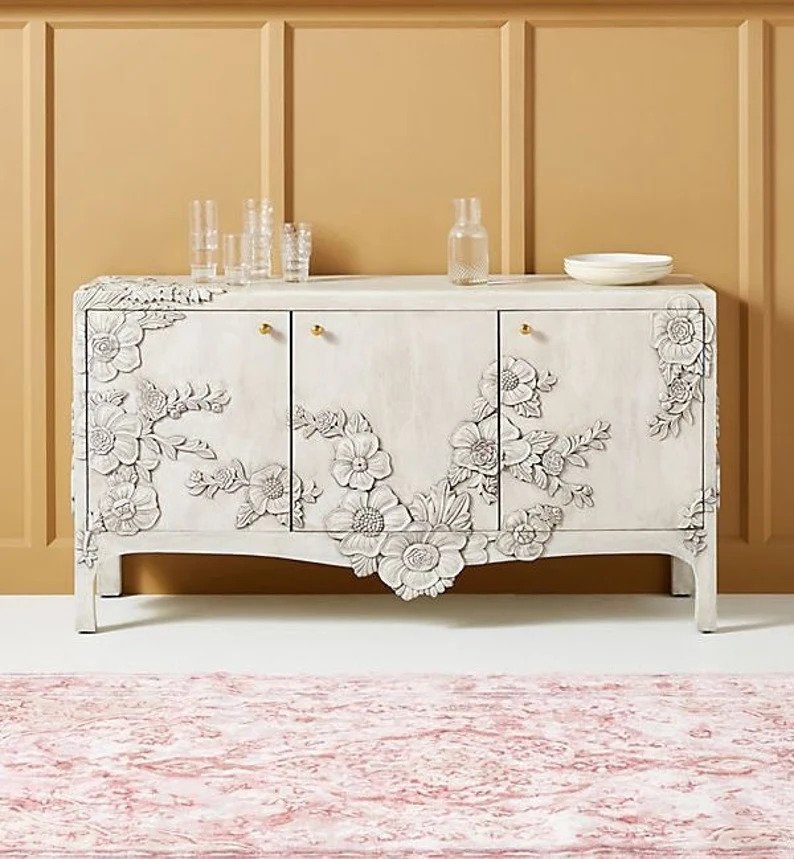 Enchantment sideboard in a white-washed finish featuring intricate, high-relief floral carvings blooming across the three cabinet doors.