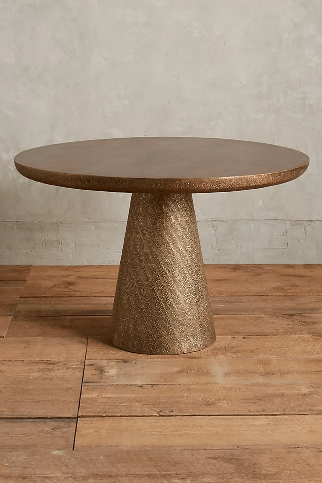 Round dining table featuring a smooth brown wood top with a bronze textured edge on a large conical hand-embossed metal base resting on wood floors.