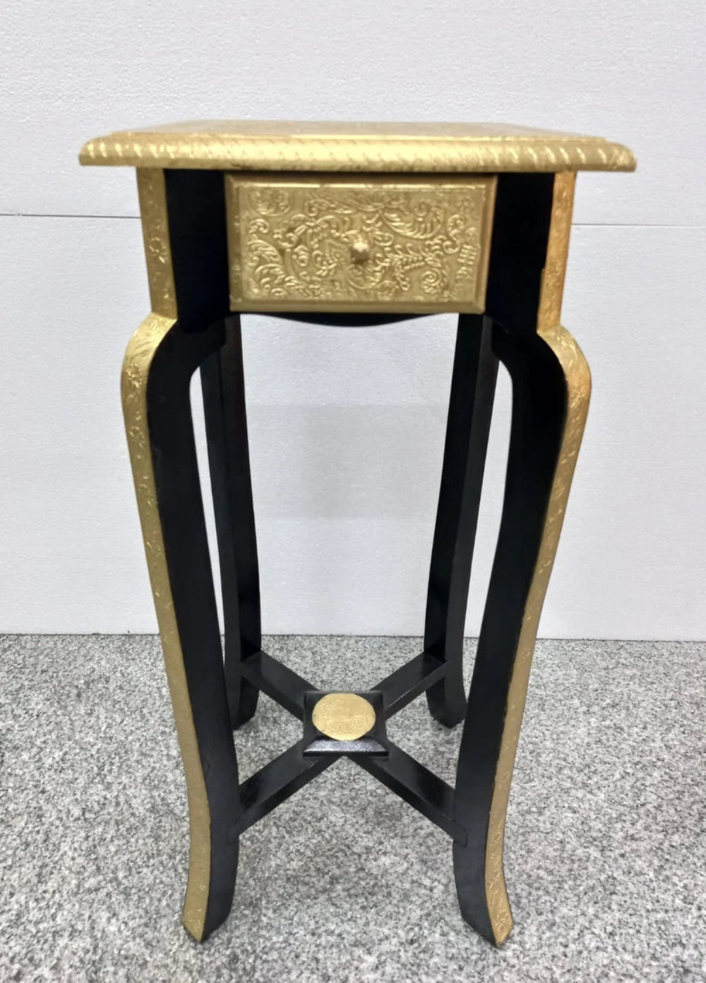 Tall traditional accent table featuring a gold foil top, an embossed gold metal floral drawer face, and black cabriole legs with an X-stretcher base.