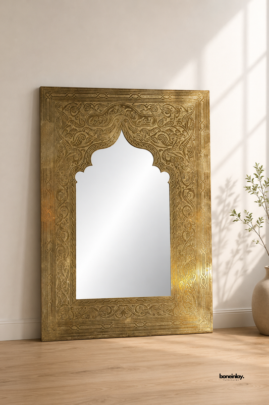 Hand-embossed brass floor mirror featuring intricate floral scrollwork and geometric border engravings with a scalloped Moroccan arch frame. Leaning against a warm neutral plaster wall in a sunlit room, this vintage brass full-length mirror brings luxurious bohemian elegance and statement artisan craftsmanship to modern interiors.
