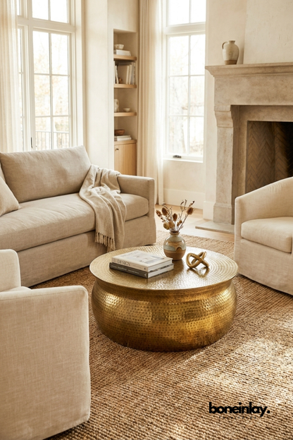 Round hammered brass drum coffee table in a warm neutral living room with a beige sofa, jute rug, and fireplace by Boneinlay Furnitures