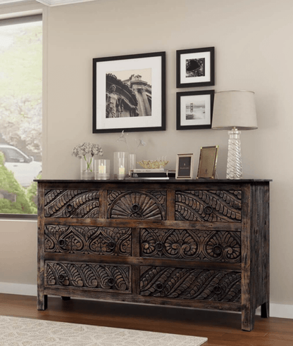 A seven-drawer wood bureau with carved floral fronts and ring pulls, styled against a wall in a living room.