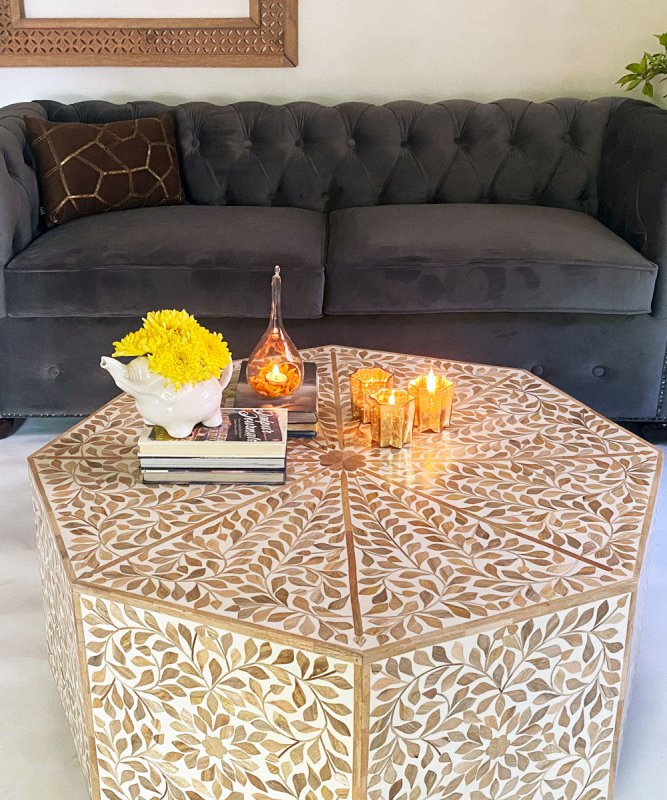 Artisan octagonal bone inlay coffee table with leaf pattern in beige and gold, styled in dark living room with tufted sofa, candles, and yellow marigold flowers from above