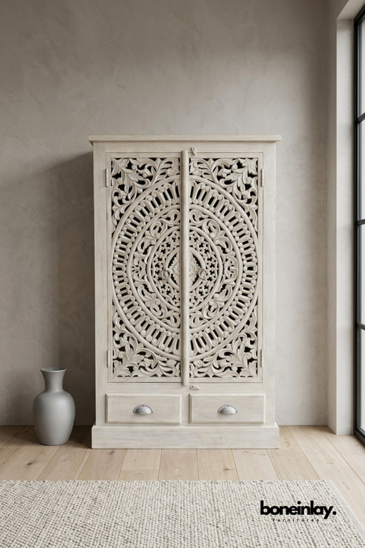 White carved wood wardrobe with a large circular mandala door pattern.
Detailed radiating carving covers the full height of both doors.
Two bottom drawers with cup pulls add extra storage space.
Styled beside a matte grey vase in a softly lit room.