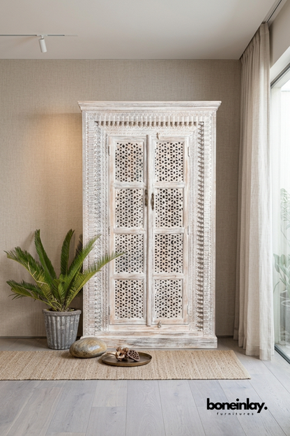 Whitewashed carved wood armoire with detailed lattice-paneled doors.
Ornate carved trim frames the top, sides, and door panels.
Four-panel star lattice design adds intricate handcrafted detail.
Styled beside a potted palm and woven jute rug.