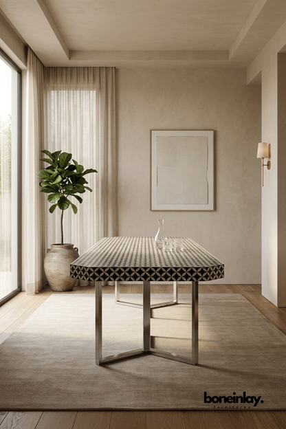 Rectangular dining table with a black and white bone inlay top.
Intricate geometric checkerboard pattern covers the entire surface.
Sits on a sleek stainless steel cross-frame base.
Styled in a minimalist dining space beside a large potted fig tree.