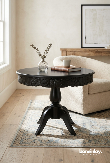 Matte black round pedestal side table with an intricate, deep-relief hand-carved floral apron, standing on a sturdy three-legged base.
