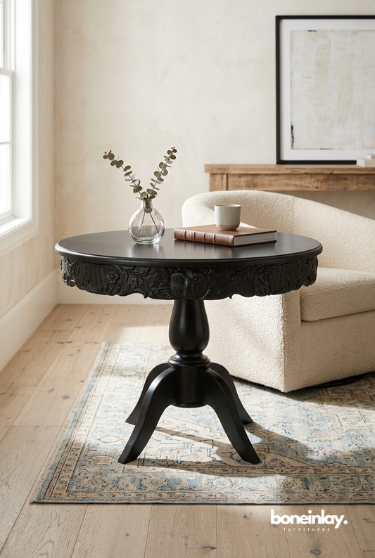 Matte black round pedestal side table with an intricate, deep-relief hand-carved floral apron, standing on a sturdy three-legged base.