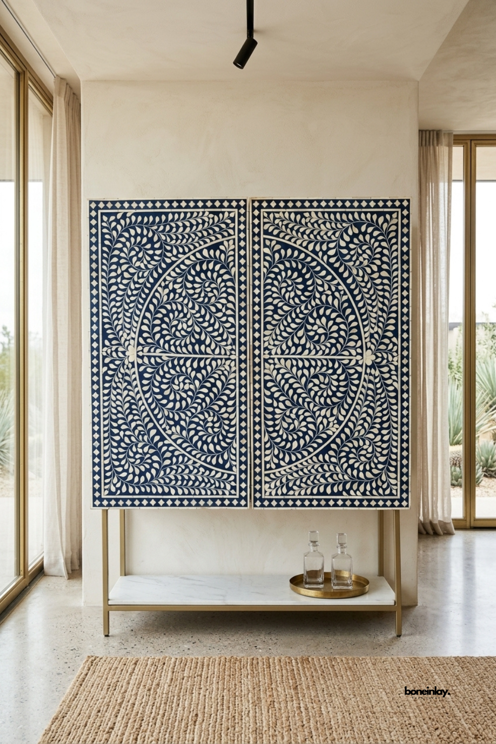 Handmade bone inlay high bar cabinet featuring a grand circular floral vine motif in crisp white against deep navy blue resin, elevated on a brushed brass metal base with an open marble bottom shelf. Styled in an airy modern living room with floor-to-ceiling windows, this artisan bar cabinet brings dramatic elegance and statement luxury.