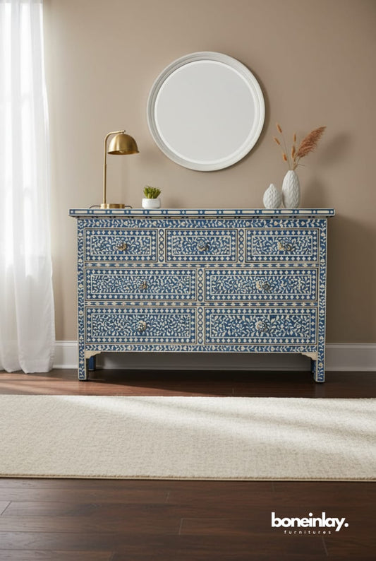 Handmade blue and white floral bone inlay seven-drawer dresser displayed in a styled living space with a round mirror, brass lamp, and decorative vases. Features intricate vine and blossom inlay pattern, floral drawer knobs, and bracket feet — artisan-crafted luxury storage furniture from Rajasthan, India.