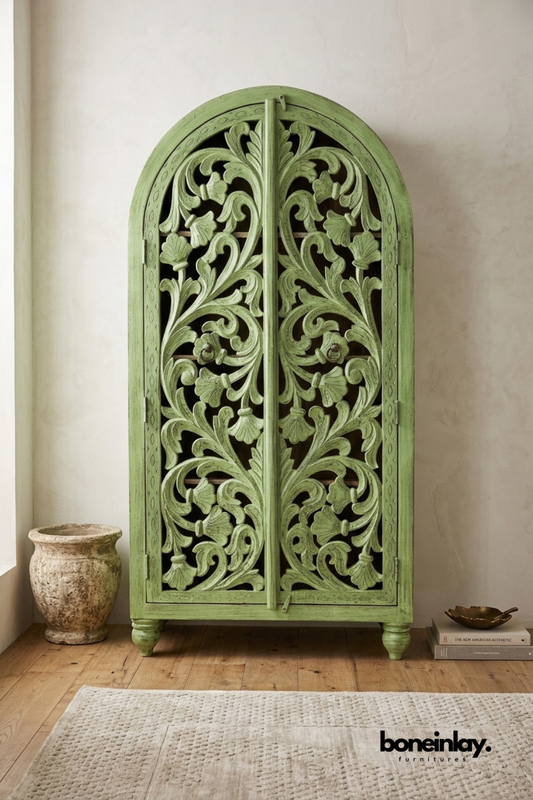 Green arched wood armoire with an intricate carved floral lattice front.
Rounded archway top features flowing vine and leaf scrollwork.
Distressed sage green finish gives it a soft, vintage character.
Styled beside a weathered stone urn on a wood floor.