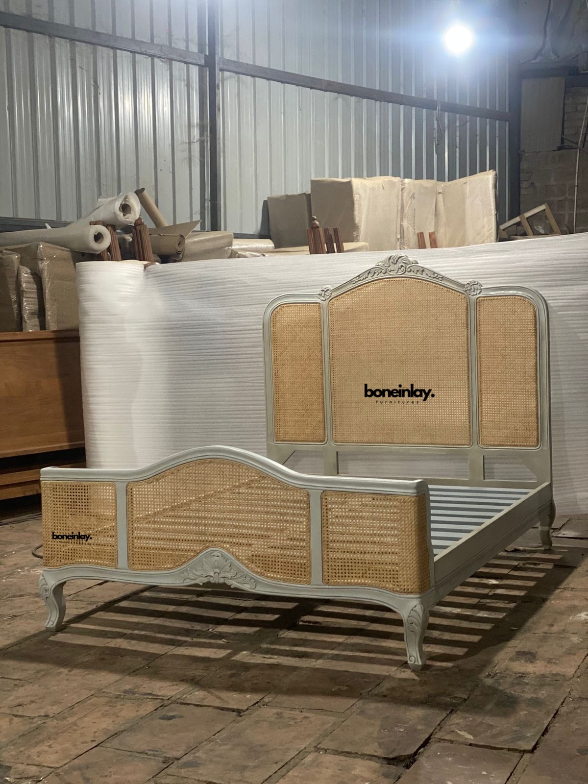 This image features a hand-carved French Provincial bed frame in a soft grey finish, characterized by its three-panel headboard and footboard. Each panel is inset with natural honey-toned cane webbing, providing a warm contrast to the carved wood frame. The design includes elegant cabriole legs and ornate floral crests, blending traditional European charm with artisanal craftsmanship.
