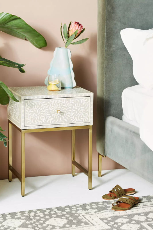 Grey bone inlay floral bedside table with single drawer and brass legs styled beside a bed with tropical plant and vase, perfect for a bohemian luxury bedroom interior.