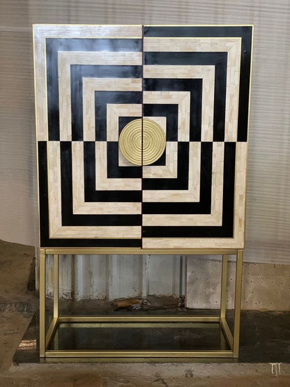 Bone inlay bar cabinet with black and white concentric square geometric pattern, brass circular door knob and gold powder-coated metal frame stand on workshop floor
