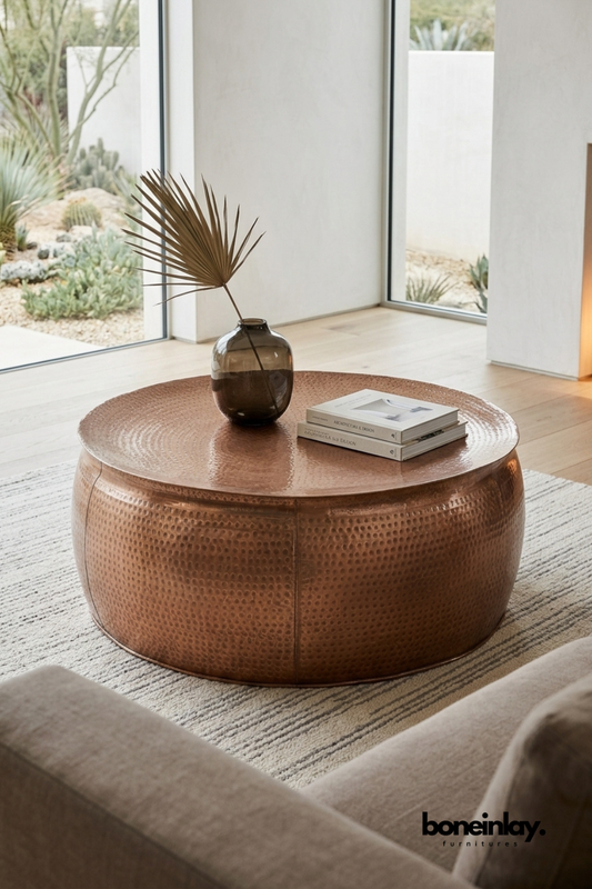 Round hammered copper coffee table featuring a rich warm patina and rounded drum silhouette. Textured hammered surface catches the light for artisanal handcrafted appeal. Styled with a smoked glass vase, dried palm frond, and stacked design books on a striped jute rug. A statement coffee table bringing warm metallic tone to modern and desert-inspired living rooms.