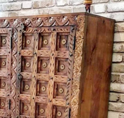 Carved panel storage cabinet in rustic wood, edge detail view showing leafy trim, recessed door panels and a ring pull.
