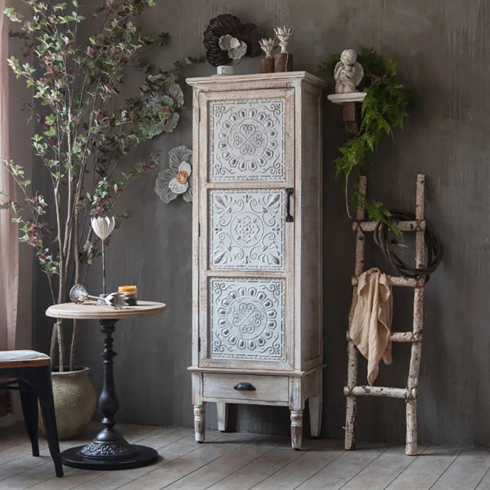 Carved wood storage cabinet with a pale floral door and lower drawer, styled in a living room with dark walls.