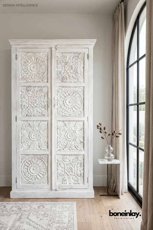 Ivory whitewashed wardrobe featuring eight hand-carved floral medallion and scroll panels across tall double doors. Classic crown molding top and brass hardware complete its traditional silhouette. Styled beside a marble side table with a glass bud vase near an arched window. A grand statement wardrobe for bedrooms, dressing rooms, and entryway storage.