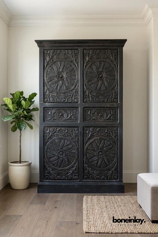 Black carved wood wardrobe with four sunburst medallion door panels.
Detailed floral and radiating carving decorates each panel section.
Crown molding top and base trim complete the traditional silhouette.
Styled beside a fiddle leaf fig plant in a light living room.