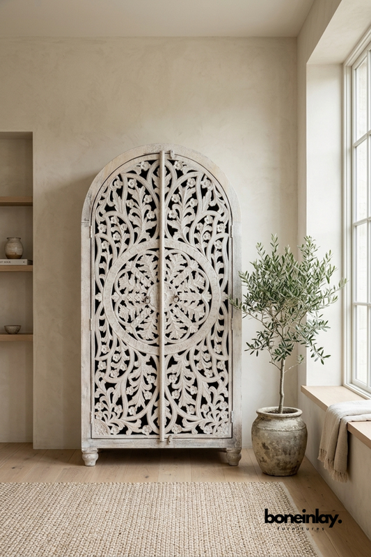 Whitewashed arched wood wardrobe with a large circular carved design.
Dense floral vine carving spans the full height of both doors.
Rounded archway top and turned feet complete the ornate silhouette.
Styled beside an olive tree near a sunlit window seat.
