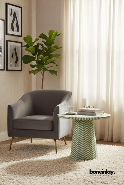 A round sage green and white chevron bone inlay side table with a pedestal base, styled next to a grey velvet armchair on a plush cream rug.