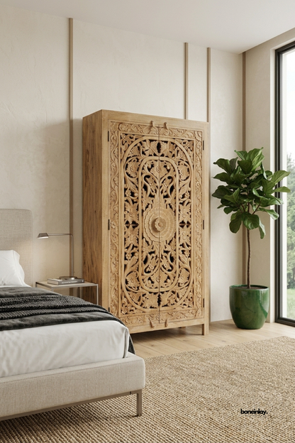 Natural mango wood wardrobe featuring a bold central floral rosette surrounded by dense scrollwork and vine carvings. Tall arched silhouette with tapered legs adds architectural presence to the bedroom setting. Styled beside a fiddle leaf fig and a linen upholstered bed with a bedside lamp. A striking statement wardrobe for bedrooms and dressing rooms.