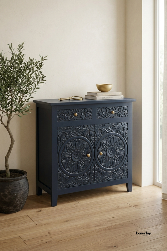 Navy blue carved wood accent cabinet featuring a bold double floral medallion pattern across two doors and a top drawer. Brass knob hardware adds refined contrast against the rich painted finish. Styled with a brass bowl, stacked books, and a vintage magnifying glass beside an olive tree. A striking statement cabinet for entryways and living rooms.