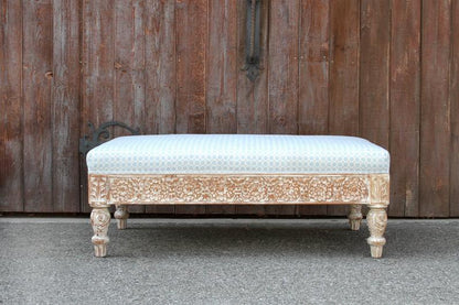 Floral carved wood ottoman in rustic finish, front view showing a patterned upholstered seat and turned wooden legs.