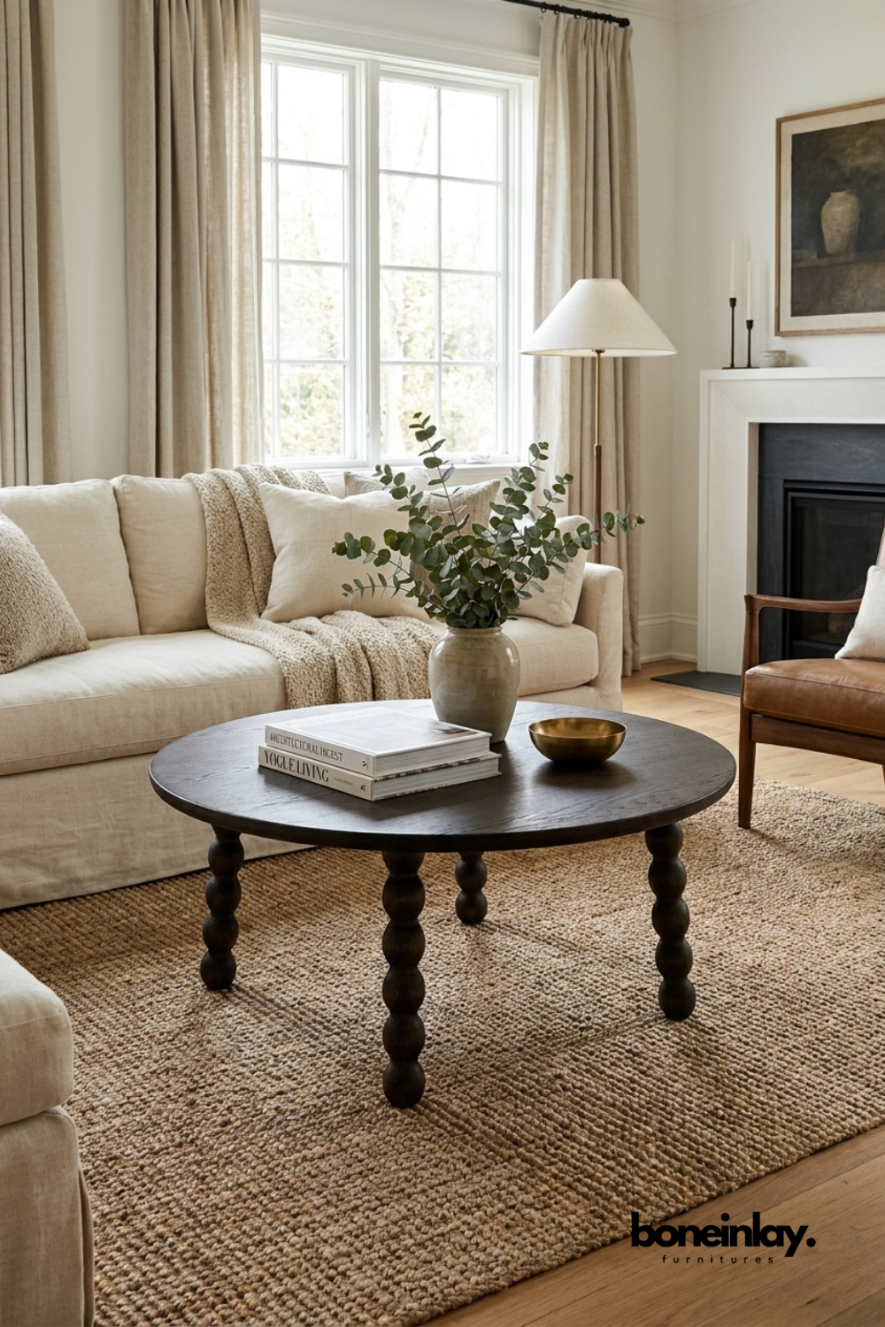 Modern living room featuring a dark wood round coffee table with turned legs, beige linen sofa, textured jute rug, and large windows with neutral curtains.