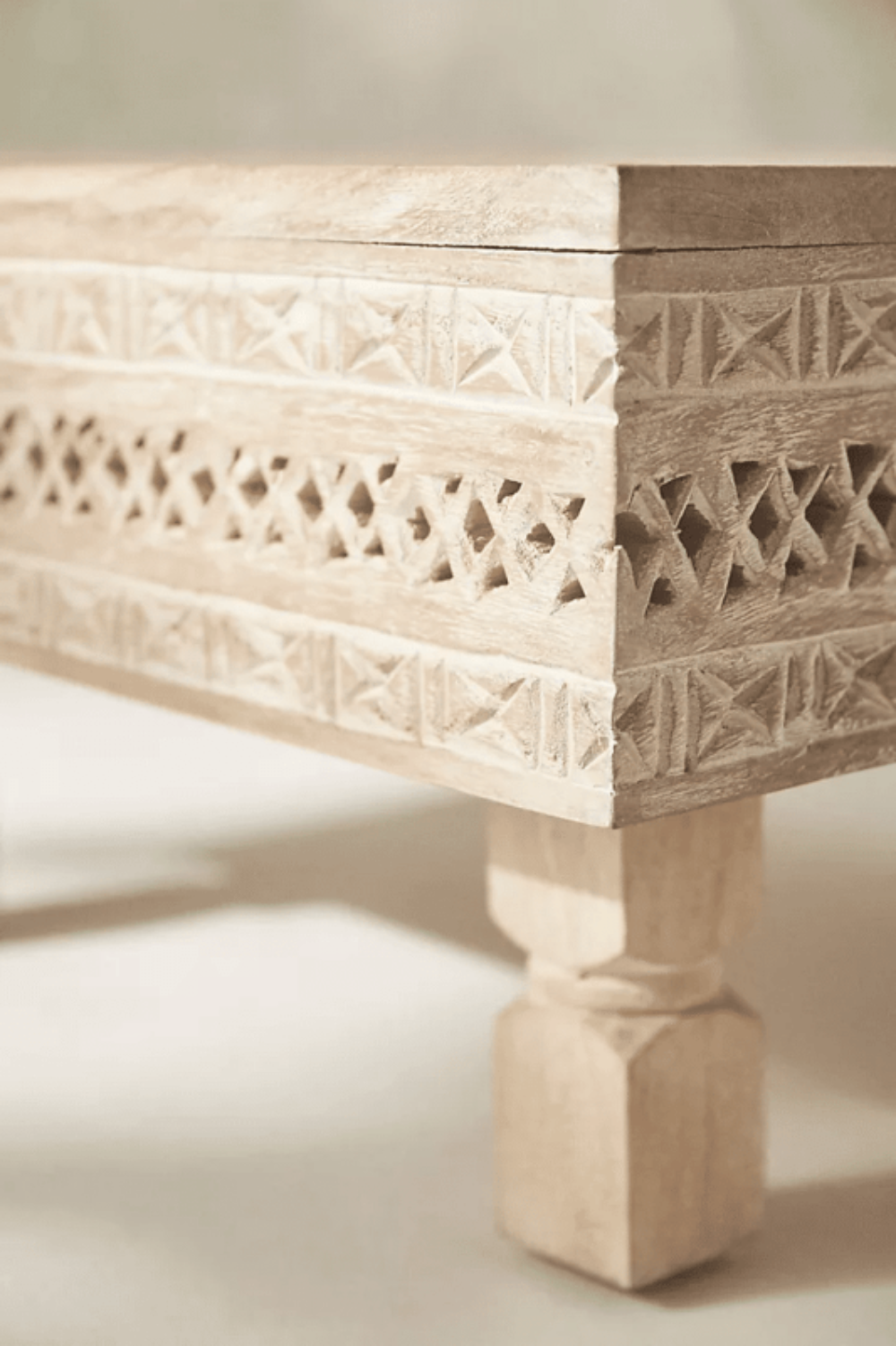 Geometric carved wood cocktail table in a light sand finish, shown in a corner detail view of its apron and leg.