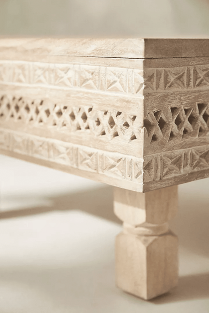 Geometric carved wood cocktail table in a light sand finish, shown in a corner detail view of its apron and leg.