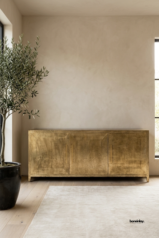 Handcrafted brass sheet-clad sideboard console featuring three framed geometric panel doors and sleek vertical brass pull handles. Styled in a warm minimalist room setting with textured plaster walls, an olive tree, and a neutral area rug, this luxury metallic credenza delivers sophisticated accent storage and mid-century glamour to modern interiors.