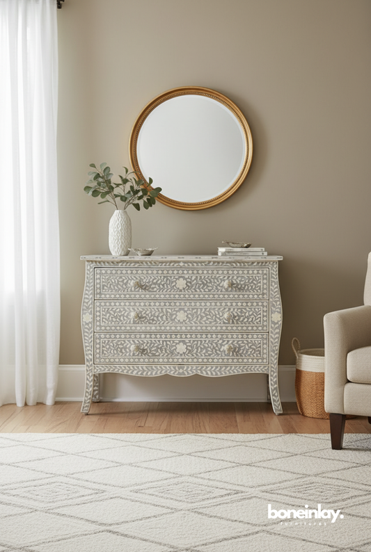Grey floral bone inlay chest of drawers with serpentine bombe body, scalloped apron, carved bone knob handles, and tapered inlaid legs, styled in a soft neutral bedroom with a gold round mirror and white vase, handcrafted artisan dresser for elegant home interiors.