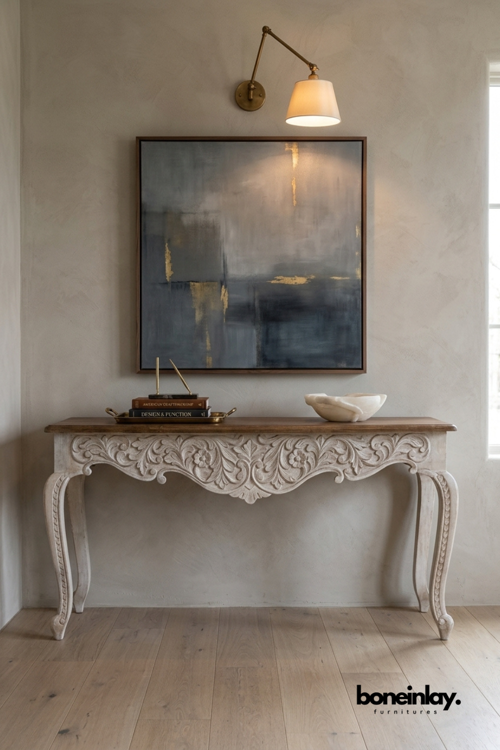 Whitewashed carved wood console table with elegant cabriole legs.
Ornate floral scroll carving decorates the apron and table skirt.
Warm wood tabletop contrasts with the distressed white-painted base.
Styled beneath a framed abstract painting and brass wall sconce.