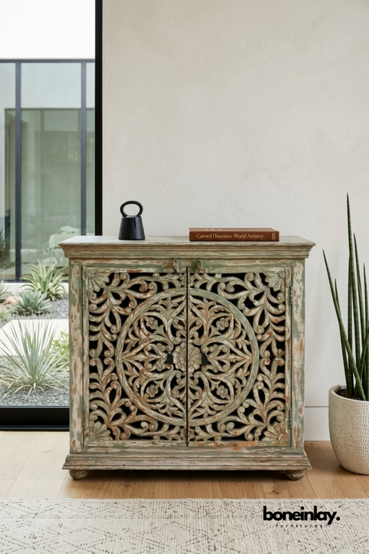 Distressed sage green carved wood accent cabinet featuring an ornate floral medallion pattern on double doors. Weathered vintage finish showcases layered rustic charm with visible wood grain. Styled with a black candle holder and leather-bound book near desert landscaping. A one-of-a-kind statement cabinet for farmhouse, vintage, and eclectic interiors.