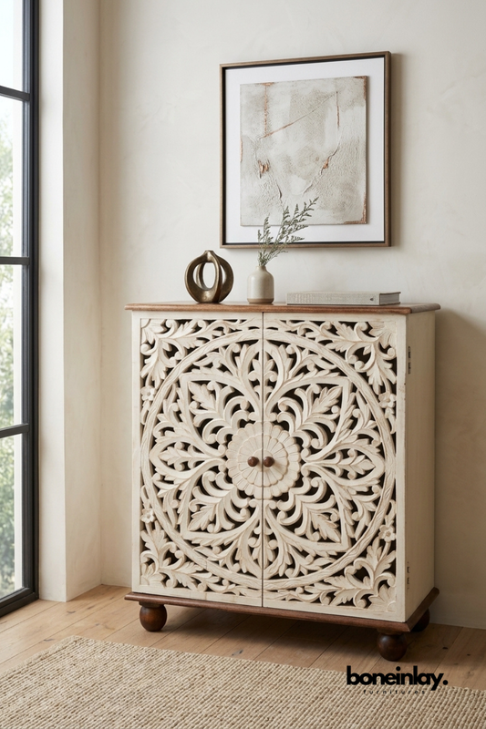 Ivory hand-carved wooden accent cabinet featuring an intricate floral medallion design on double doors. Finished with a natural wood top and round bun feet for a boho-chic look. Styled beneath framed wall art with a ceramic vase and decorative sculpture. Perfect entryway or accent cabinet for adding artisanal charm to any room.