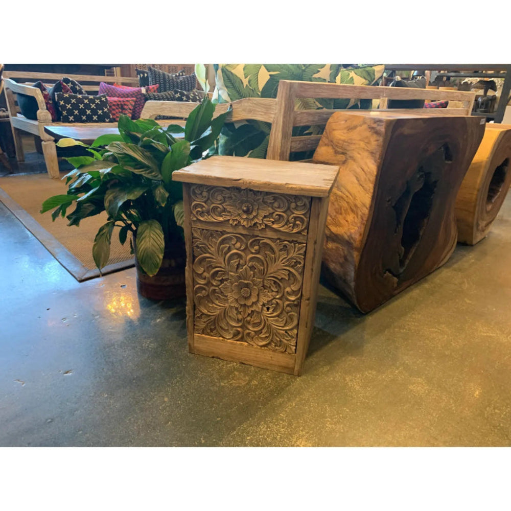 Hand-carved floral storage cabinet in solid wood, front view beside a leafy plant and stacked showroom furniture.