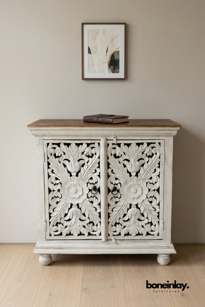 Whitewashed carved wood cabinet with a natural wood tabletop.
Floral medallion carving decorates both cabinet door fronts.
Crown molding top and round bun feet add classic detailing.
Styled below a framed abstract print in a neutral hallway.