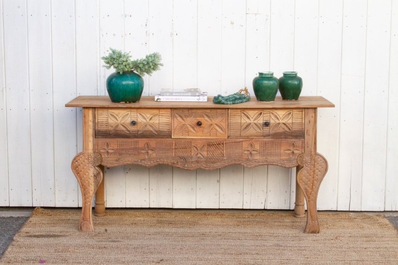 Hand-Carved Hallway Table in rustic solid wood, front view with three drawers, curved legs and plants on top.
