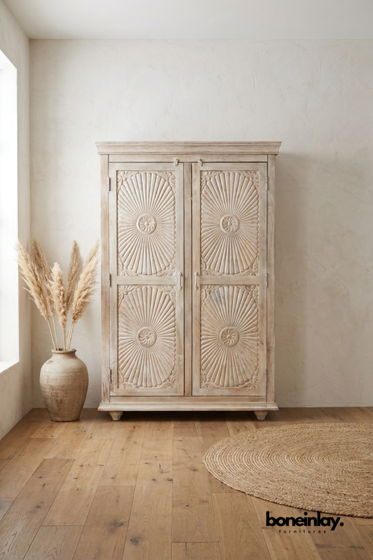 Whitewashed carved wood armoire with a bold sunburst rosette pattern.
Four carved door panels feature radiating fluted detailing.
Crown molding top and turned bun feet complete the classic silhouette.
Styled beside a stoneware vase and dried pampas grass.