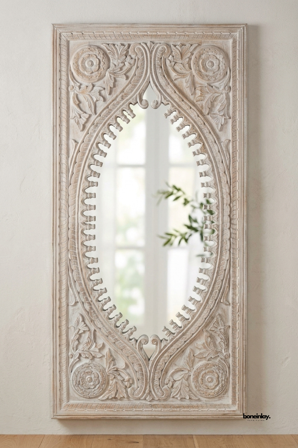 Whitewashed carved wood wall mirror featuring an ornate oval silhouette with scalloped edging and floral rosette corners. Rich acanthus leaf and scroll detailing adds traditional architectural character to the frame. Styled against a soft plaster wall with a trailing olive branch reflection. A dramatic statement mirror for entryways and formal living spaces.