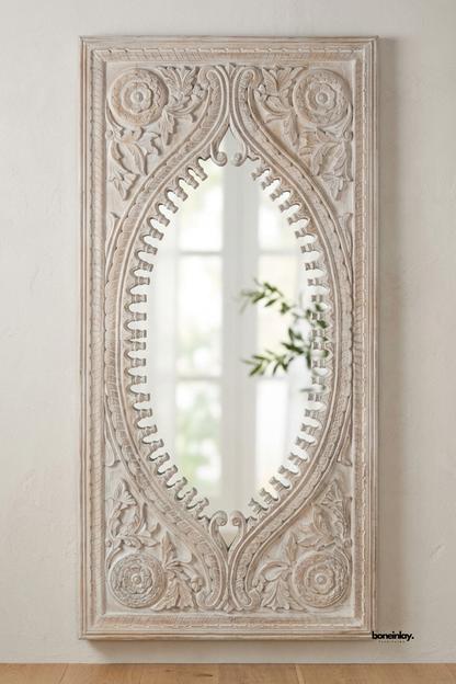 Whitewashed carved wood wall mirror featuring an ornate oval silhouette with scalloped edging and floral rosette corners. Rich acanthus leaf and scroll detailing adds traditional architectural character to the frame. Styled against a soft plaster wall with a trailing olive branch reflection. A dramatic statement mirror for entryways and formal living spaces.