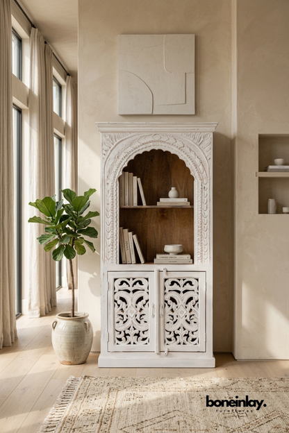 Whitewashed arched carved wood bookcase cabinet with open display shelves.
Hand-carved floral and leaf trim frames the arched top section.
Bottom half features two carved lattice cabinet doors for closed storage.
Styled beside a fiddle leaf fig plant against a warm plaster wall.