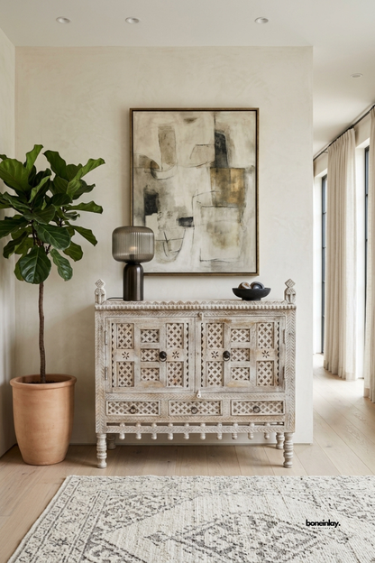 Artisan-crafted storage cabinet featuring double lattice-carved doors, three lower utility
drawers, turned spindle legs, and corner finials in an antique distressed whitewash finish.
Styled in a bright living space beside a potted fiddle-leaf fig and modern lamp,
this heirloom-style credenza provides rich textural artistry and charming organization.