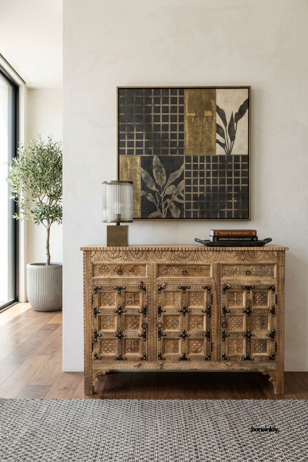 Hand-carved rustic 3-door credenza featuring paneled geometric relief carvings,
three upper utility drawers, and traditional dark iron strap and hinge accents.
Styled in an elegant foyer beneath modern mixed-media art with an olive tree
and fluted glass lamp, this artisan sideboard brings heritage warmth and statement storage.